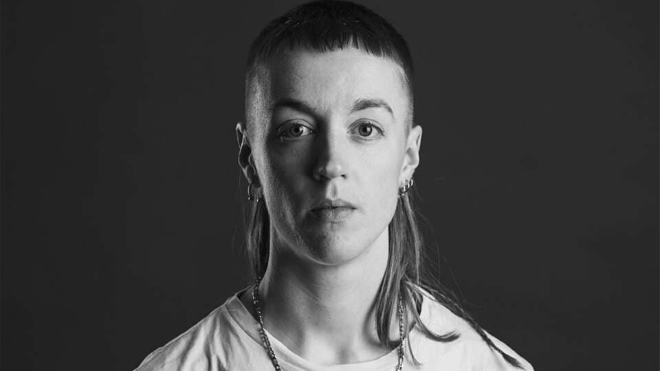 A person with short bangs and long hair at the back, wearing earrings and a necklace, is looking directly at the camera in a black and white portrait.