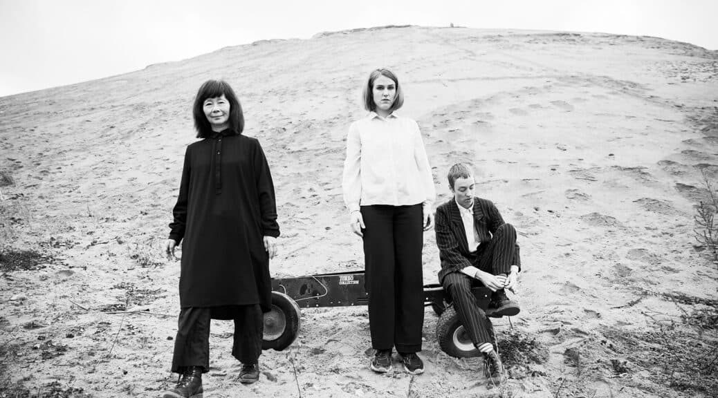 Three people stand on a sandy landscape, with one sitting on a small wagon.