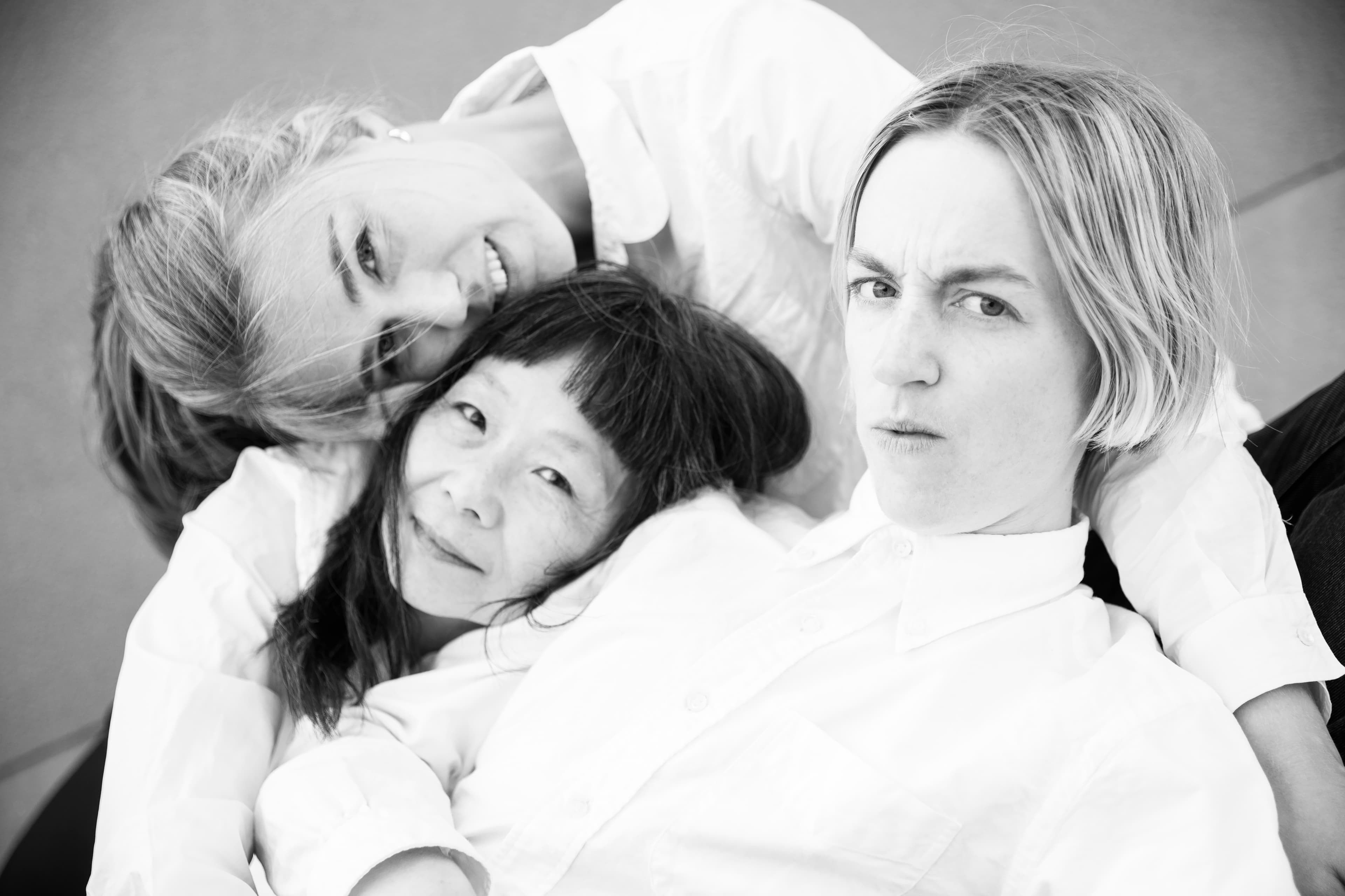Three people wearing white shirts are lying together closely, with one person looking directly at the camera.