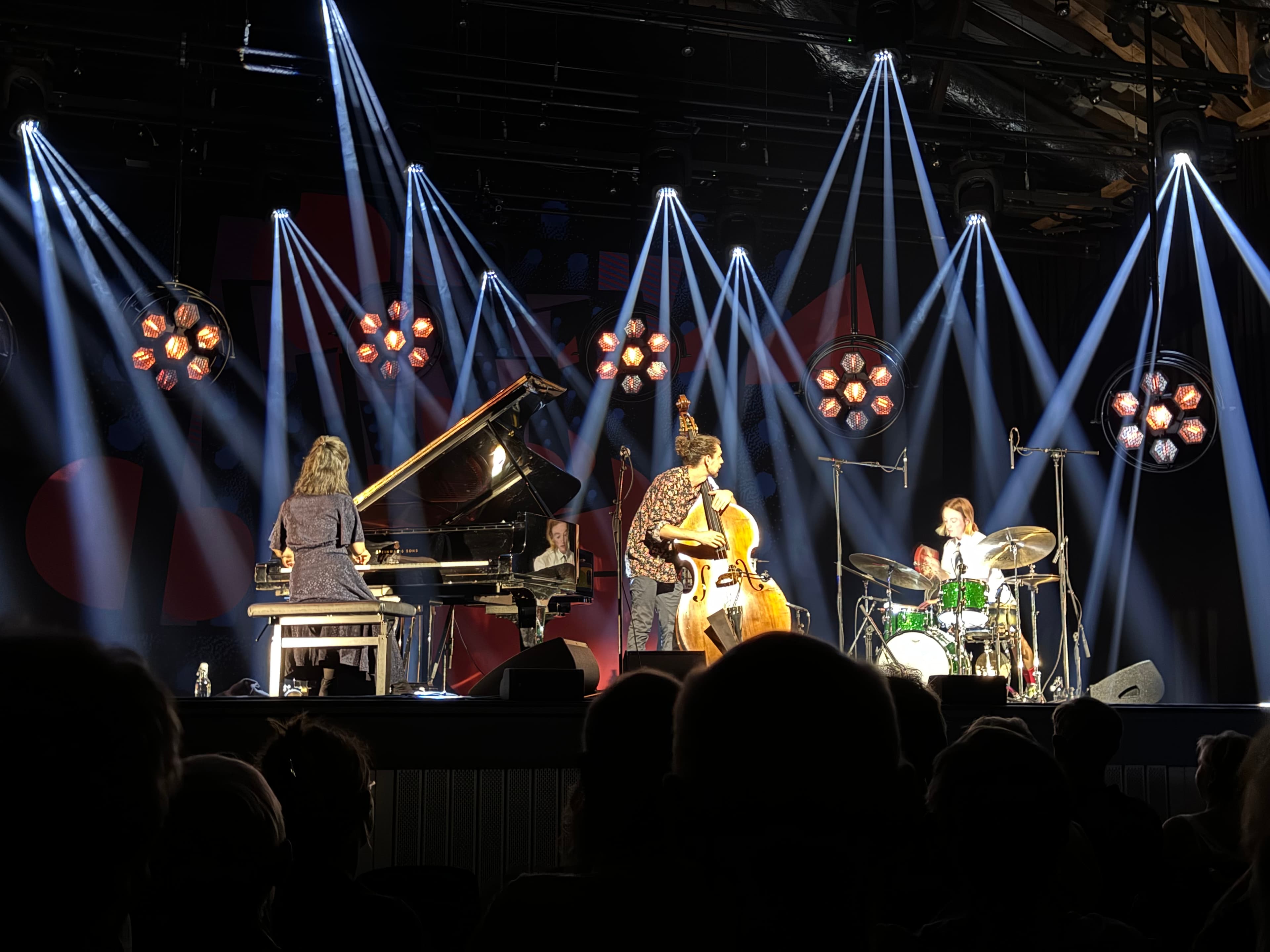 Three musicians perform on stage with a pianist, a double bassist, and a drummer, under bright stage lights.